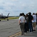 A group of civilians and personnel wearing hearing protection stand along the edge of a military airfield tarmac, observing an F-35 Lightning II stealth fighter jet taxiing on the runway. The setting appears to be an organized visit or demonstration event at a military air base, with hangars visible in the background. The observers are lined up in a row, several shielding their eyes or adjusting their ear protection.