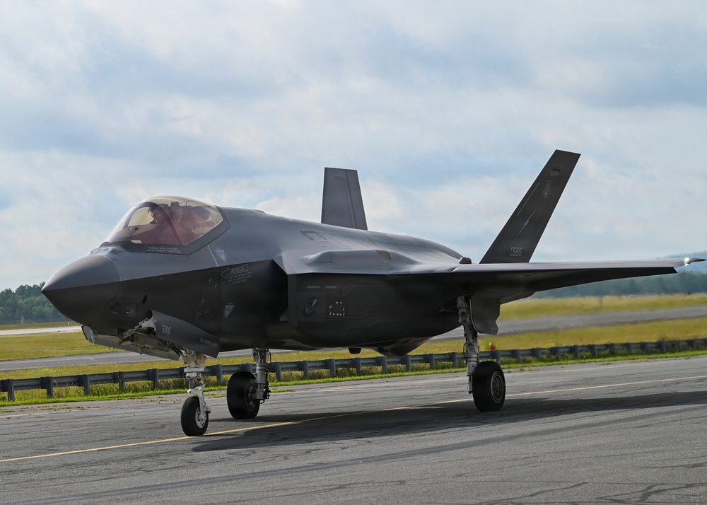 A Lockheed Martin F-35 Lightning II stealth fighter jet is taxiing on a runway apron. The aircraft is painted in a dark gray low-observable finish and has its landing gear deployed. A pilot is visible in the cockpit, and the aircraft appears to be at a military or joint-use airfield.