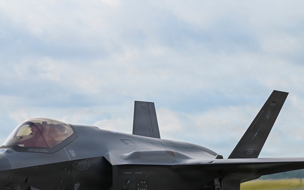 A Lockheed Martin F-35 Lightning II stealth fighter jet is taxiing on a runway apron. The aircraft is painted in a dark gray low-observable finish and has its landing gear deployed. A pilot is visible in the cockpit, and the aircraft appears to be at a military or joint-use airfield.