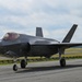A Lockheed Martin F-35 Lightning II stealth fighter jet is taxiing on a runway apron. The aircraft is painted in a dark gray low-observable finish and has its landing gear deployed. A pilot is visible in the cockpit, and the aircraft appears to be at a military or joint-use airfield.