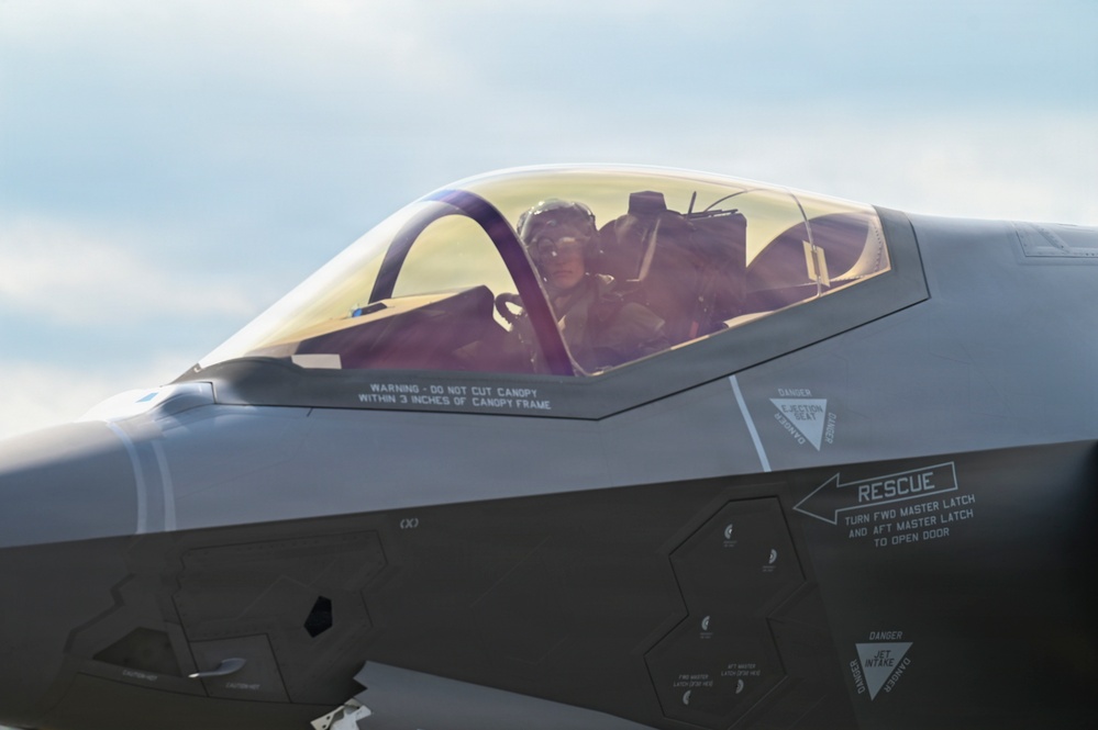 A close-up photograph of an F-35 Lightning II fighter jet cockpit with a pilot seated inside wearing a flight helmet and oxygen mask. The image captures detailed rescue and warning markings on the fuselage, including canopy cut instructions and rescue door access directions. The golden-tinted canopy reflects ambient light, giving the image a warm tone.