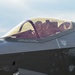 A close-up photograph of an F-35 Lightning II fighter jet cockpit with a pilot seated inside wearing a flight helmet and oxygen mask. The image captures detailed rescue and warning markings on the fuselage, including canopy cut instructions and rescue door access directions. The golden-tinted canopy reflects ambient light, giving the image a warm tone.