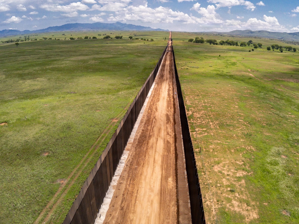 An aerial drone photograph taken from directly above a long border barrier wall stretching to the horizon across open grassland terrain. The wall consists of two parallel steel/metal fence structures with a dirt patrol road running between them. Mountains are visible in the background under a partly cloudy sky.