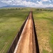An aerial drone photograph taken from directly above a long border barrier wall stretching to the horizon across open grassland terrain. The wall consists of two parallel steel/metal fence structures with a dirt patrol road running between them. Mountains are visible in the background under a partly cloudy sky.