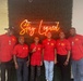 Six individuals are posing together in front of a neon sign that reads 'Stay Liquid' mounted on a green hedge wall backdrop. All six are wearing matching red polo shirts with a 'Double Time Sports Arena' logo, suggesting they are affiliated with the same organization or team. The group includes adults and two younger individuals, likely at a sports facility or business venue.