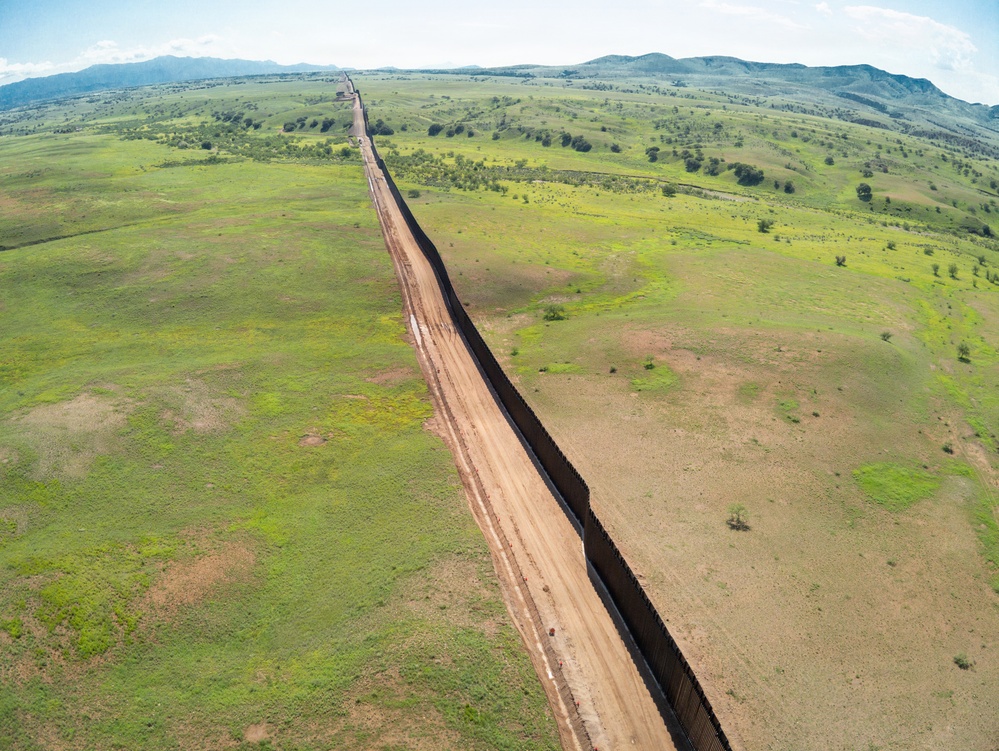 An aerial drone photograph showing a long border barrier or security fence stretching across a rural, semi-arid landscape. The dark metal fence runs diagonally through the frame, with a dirt patrol road running parallel on the left (western) side. The terrain on both sides features green grasslands, scattered shrubs, and distant mountain ranges under a partly cloudy sky.