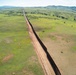 An aerial drone photograph showing a long border barrier or security fence stretching across a rural, semi-arid landscape. The dark metal fence runs diagonally through the frame, with a dirt patrol road running parallel on the left (western) side. The terrain on both sides features green grasslands, scattered shrubs, and distant mountain ranges under a partly cloudy sky.
