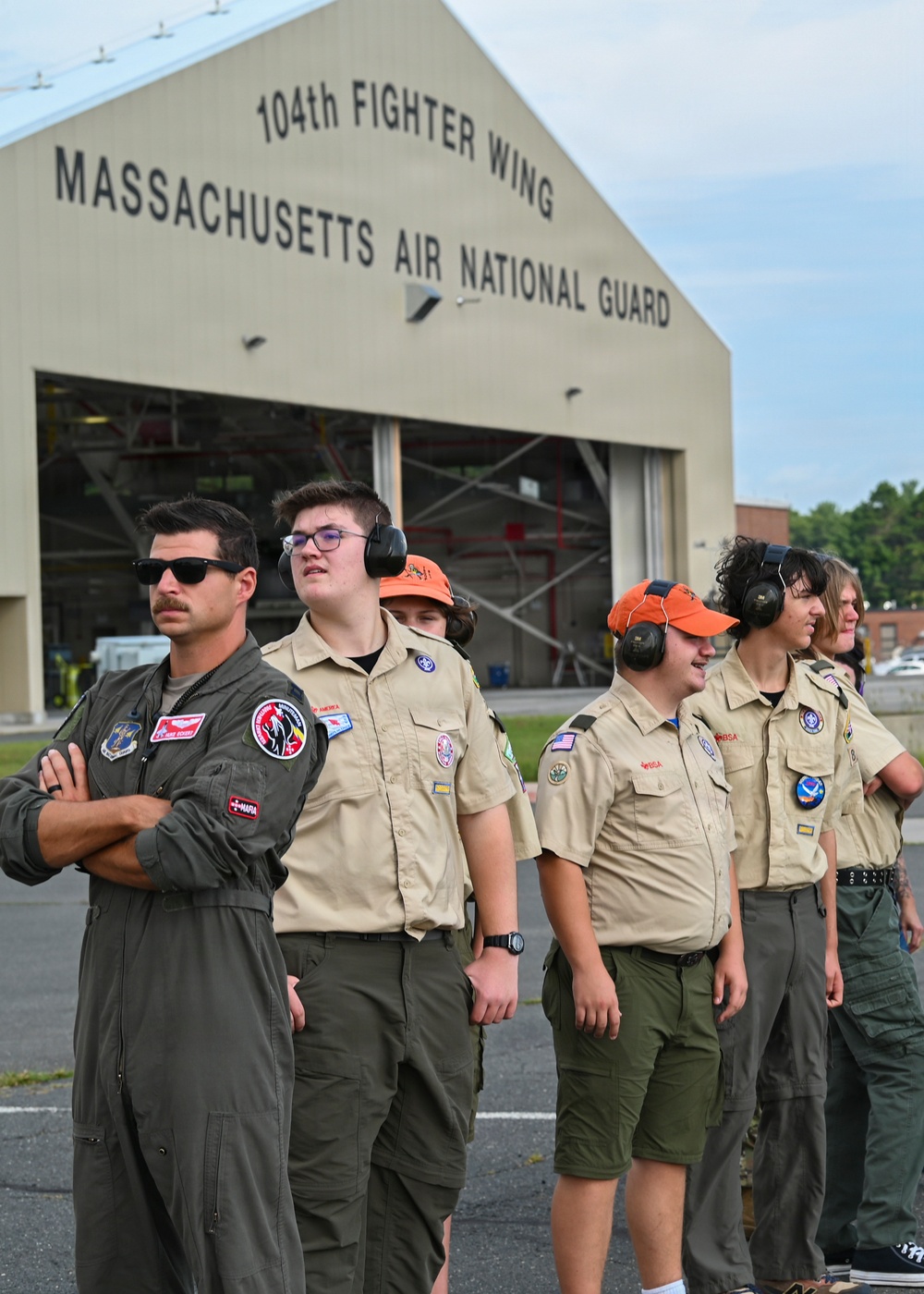 A group of Boy Scouts of America members stand on the tarmac at the 104th Fighter Wing, Massachusetts Air National Guard base. They are accompanied by a military pilot in an olive flight suit with patches and sunglasses. Several scouts are wearing hearing protection ear muffs and orange caps, suggesting they are observing flight operations.