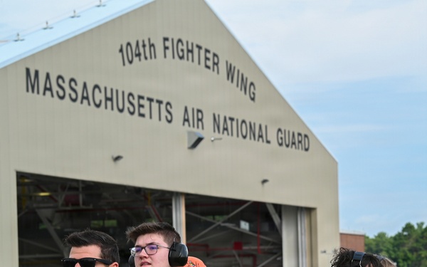 A group of Boy Scouts of America members stand on the tarmac at the 104th Fighter Wing, Massachusetts Air National Guard base. They are accompanied by a military pilot in an olive flight suit with patches and sunglasses. Several scouts are wearing hearing protection ear muffs and orange caps, suggesting they are observing flight operations.