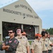 A group of Boy Scouts of America members stand on the tarmac at the 104th Fighter Wing, Massachusetts Air National Guard base. They are accompanied by a military pilot in an olive flight suit with patches and sunglasses. Several scouts are wearing hearing protection ear muffs and orange caps, suggesting they are observing flight operations.