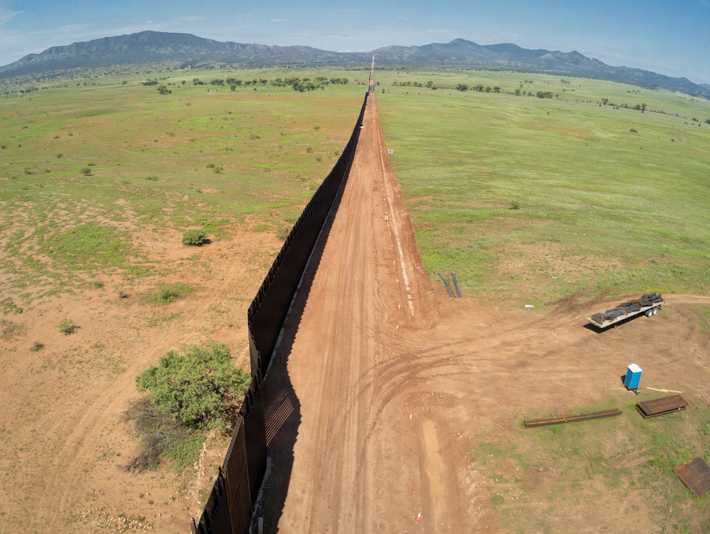 An aerial photograph showing a long border barrier/fence stretching to the horizon across open grassland terrain. The dark metal fence runs in a straight line dividing two landscapes, with a dirt patrol road running parallel on one side. Construction equipment and materials are visible near the foreground, suggesting active construction or maintenance work.