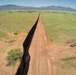 An aerial photograph showing a long border barrier/fence stretching to the horizon across open grassland terrain. The dark metal fence runs in a straight line dividing two landscapes, with a dirt patrol road running parallel on one side. Construction equipment and materials are visible near the foreground, suggesting active construction or maintenance work.