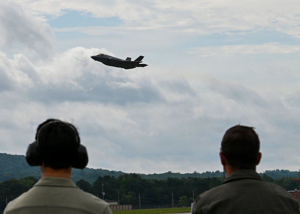 Two individuals, viewed from behind, observe a military jet aircraft in flight over a wooded airfield. The aircraft, identifiable by its silhouette as an F-35 Lightning II, is captured mid-flight against a partly cloudy sky. One observer is wearing hearing protection ear muffs, suggesting proximity to loud aircraft operations.