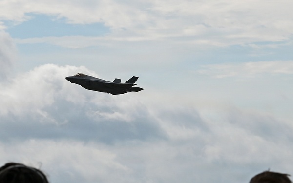 Two individuals, viewed from behind, observe a military jet aircraft in flight over a wooded airfield. The aircraft, identifiable by its silhouette as an F-35 Lightning II, is captured mid-flight against a partly cloudy sky. One observer is wearing hearing protection ear muffs, suggesting proximity to loud aircraft operations.