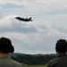 Two individuals, viewed from behind, observe a military jet aircraft in flight over a wooded airfield. The aircraft, identifiable by its silhouette as an F-35 Lightning II, is captured mid-flight against a partly cloudy sky. One observer is wearing hearing protection ear muffs, suggesting proximity to loud aircraft operations.