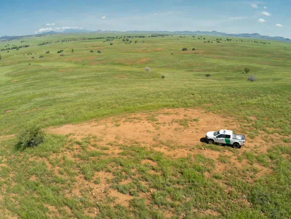 An aerial drone photograph captures a white government pickup truck parked on a dirt clearing in a vast, rolling green grassland. The vehicle appears to be a U.S. Border Patrol truck, identifiable by its white color and green stripe markings. The landscape features expansive open rangeland with scattered trees, red soil patches, and distant mountain ranges under a partly cloudy sky.