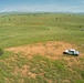 An aerial drone photograph captures a white government pickup truck parked on a dirt clearing in a vast, rolling green grassland. The vehicle appears to be a U.S. Border Patrol truck, identifiable by its white color and green stripe markings. The landscape features expansive open rangeland with scattered trees, red soil patches, and distant mountain ranges under a partly cloudy sky.