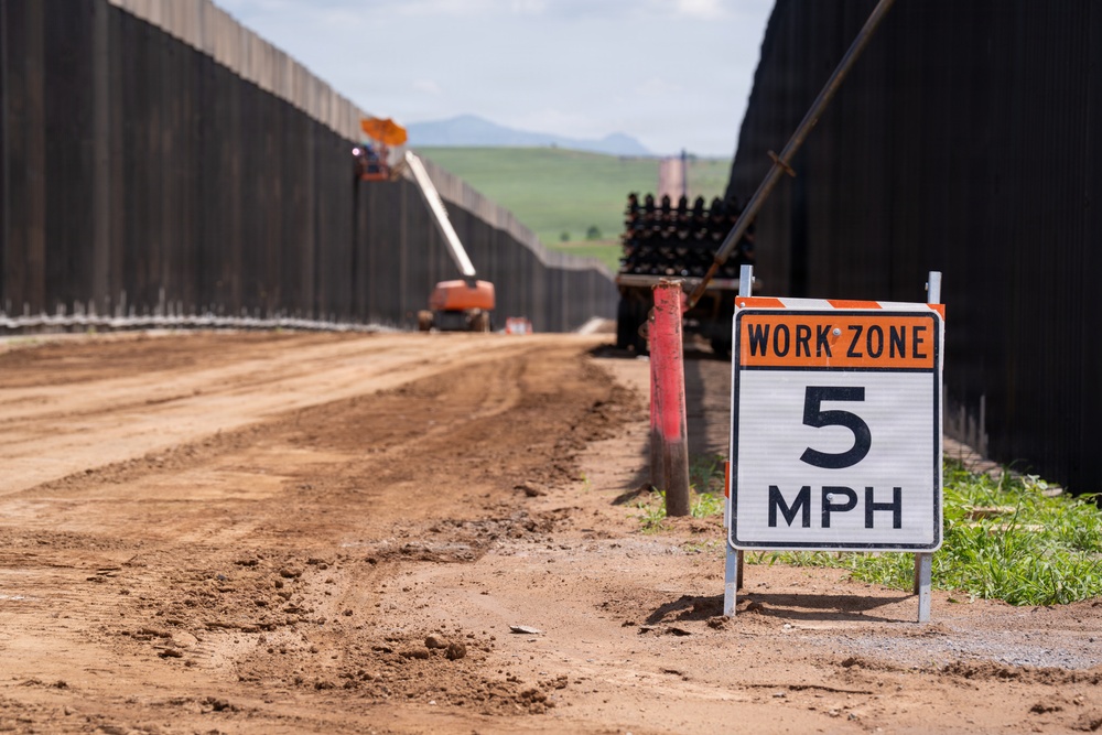 A construction work zone is visible between two tall dark metal barrier walls, with a dirt road running between them. A 'Work Zone 5 MPH' speed limit sign is prominently displayed in the foreground. Construction equipment, including an orange boom lift, is visible in the background actively working on the barriers.