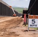 A construction work zone is visible between two tall dark metal barrier walls, with a dirt road running between them. A 'Work Zone 5 MPH' speed limit sign is prominently displayed in the foreground. Construction equipment, including an orange boom lift, is visible in the background actively working on the barriers.