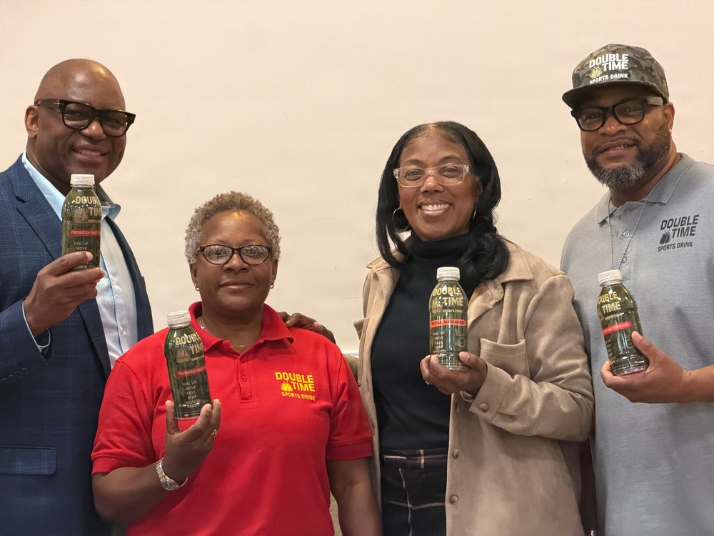 Four adults are posing together while holding bottles of 'Double Time Sports Drink.' Two of the individuals are wearing branded Double Time Sports Drink apparel, suggesting they may be associated with the brand. The group appears to be at a promotional or product showcase event.