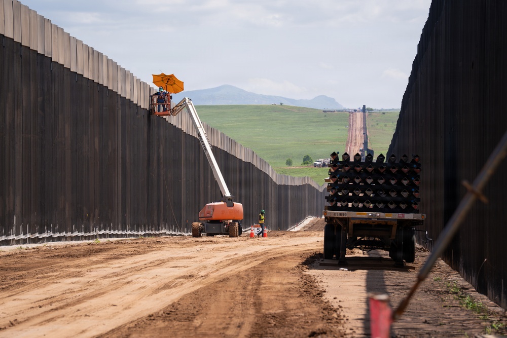 The image shows an active large-scale border wall construction site, with tall dark steel bollard panels extending into the distance on both sides of a dirt service road. Construction equipment including a boom lift with an orange umbrella shade and a flatbed truck loaded with steel bollard sections are visible. The setting appears to be a rural, semi-arid landscape with green hills and mountains visible in the background.