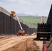 The image shows an active large-scale border wall construction site, with tall dark steel bollard panels extending into the distance on both sides of a dirt service road. Construction equipment including a boom lift with an orange umbrella shade and a flatbed truck loaded with steel bollard sections are visible. The setting appears to be a rural, semi-arid landscape with green hills and mountains visible in the background.