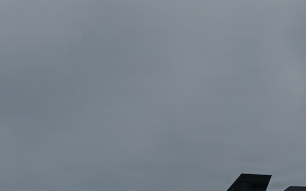 A single military stealth fighter jet is captured in low-level flight against an overcast gray sky. The aircraft displays characteristic stealth design features including angular surfaces, a single-seat cockpit, and canted twin tail fins. The image was taken in dim, flat lighting conditions which reduces overall contrast and detail.