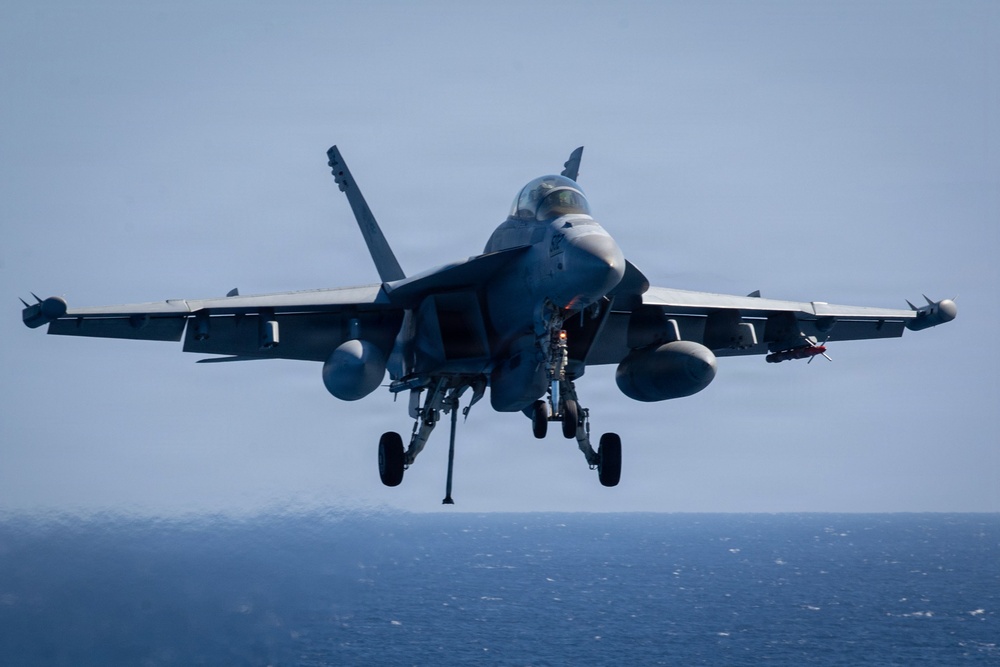 A carrier-based twin-engine jet fighter is captured in a low-altitude approach, likely preparing to land on an aircraft carrier over open ocean. The aircraft has its landing gear fully deployed and is carrying external ordnance and fuel systems. The image is taken from a forward-facing perspective, providing a clear frontal view of the aircraft.