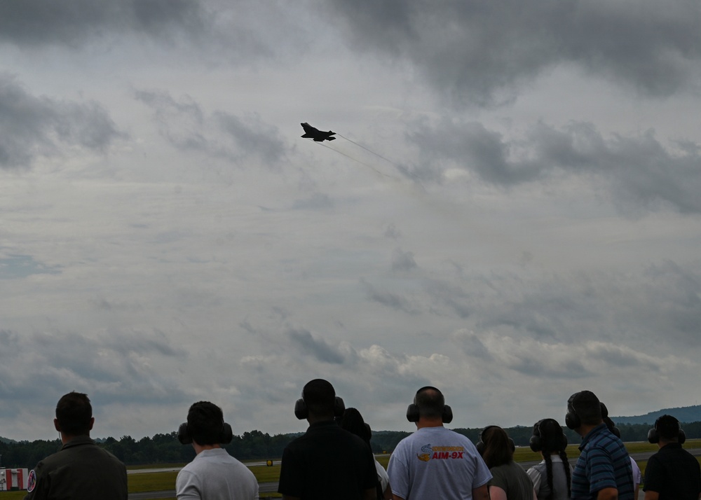 A crowd of spectators at an airshow watches a military jet aircraft performing a high-speed pass against an overcast sky. Several observers are wearing hearing protection ear muffs, and one person is wearing a shirt with 'AIM-9X' printed on it, suggesting a military aviation context. The aircraft leaves a condensation trail as it climbs steeply.