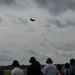 A crowd of spectators at an airshow watches a military jet aircraft performing a high-speed pass against an overcast sky. Several observers are wearing hearing protection ear muffs, and one person is wearing a shirt with 'AIM-9X' printed on it, suggesting a military aviation context. The aircraft leaves a condensation trail as it climbs steeply.