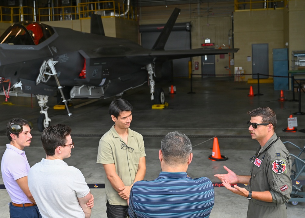 A military pilot in an olive flight suit is briefing or speaking to a small group of four civilians inside an aircraft hangar. Behind the group, an F-35 Lightning II stealth fighter jet is parked on the hangar floor. The setting appears to be an official tour or informational visit to a military air base.