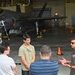 A military pilot in an olive flight suit is briefing or speaking to a small group of four civilians inside an aircraft hangar. Behind the group, an F-35 Lightning II stealth fighter jet is parked on the hangar floor. The setting appears to be an official tour or informational visit to a military air base.
