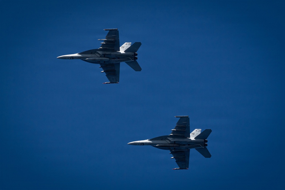 Two military jet fighters are captured in flight against a clear, deep blue sky. Both aircraft appear to be F/A-18 Hornet or Super Hornet variants, flying in a loose formation with one aircraft inverted or banking above the other. The image is taken from a ground-level perspective looking upward, providing a clear view of the underside of both aircraft.