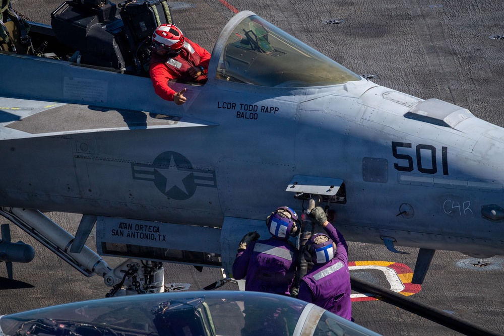 A U.S. Navy carrier-based aircraft (numbered 501) is shown on a flight deck with crew members performing pre-flight or maintenance activities. The pilot, wearing a red helmet and red flight suit, is seated in the cockpit giving a thumbs-up gesture. Two deck crew members in purple jerseys are working on the aircraft's fuselage.