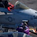 A U.S. Navy carrier-based aircraft (numbered 501) is shown on a flight deck with crew members performing pre-flight or maintenance activities. The pilot, wearing a red helmet and red flight suit, is seated in the cockpit giving a thumbs-up gesture. Two deck crew members in purple jerseys are working on the aircraft's fuselage.