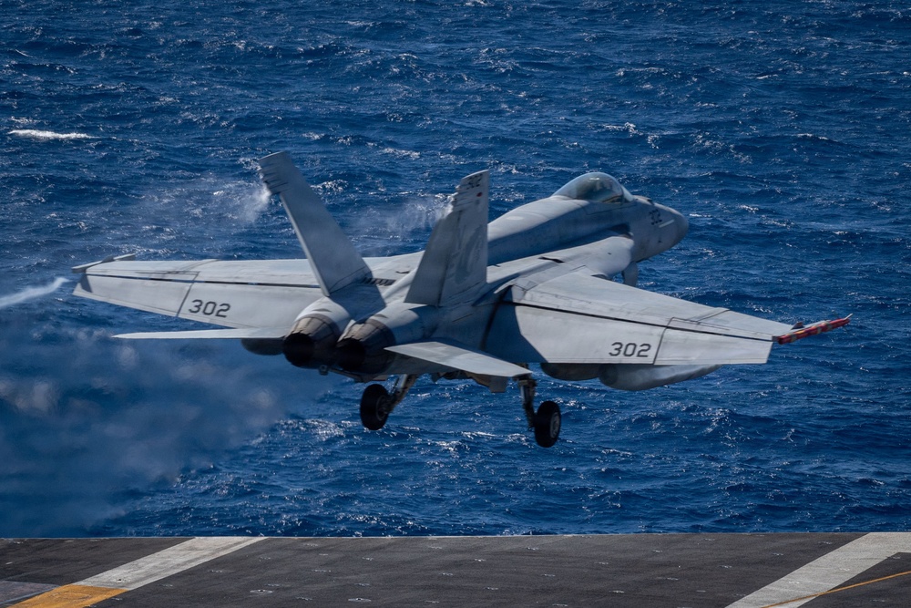 A U.S. Navy carrier-based jet aircraft marked '302' is captured at the moment of catapult launch from an aircraft carrier flight deck over open ocean. The aircraft is airborne with landing gear still extended, and visible steam or exhaust trails behind it from the catapult system. The deep blue ocean serves as the backdrop, indicating operations far from shore.