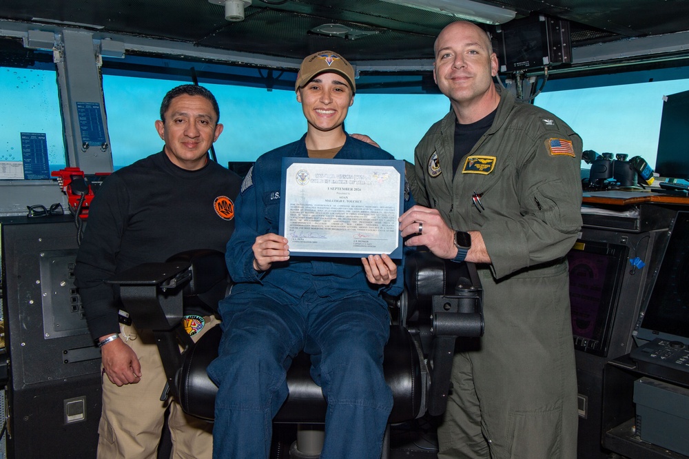 Three military personnel pose for a photo aboard what appears to be a naval vessel's bridge or command area. The central figure, seated in a command chair, is holding a certificate dated 1 September 2026, suggesting a promotion or award ceremony. The individuals are wearing different military uniforms indicating possible inter-service or mixed-role personnel.