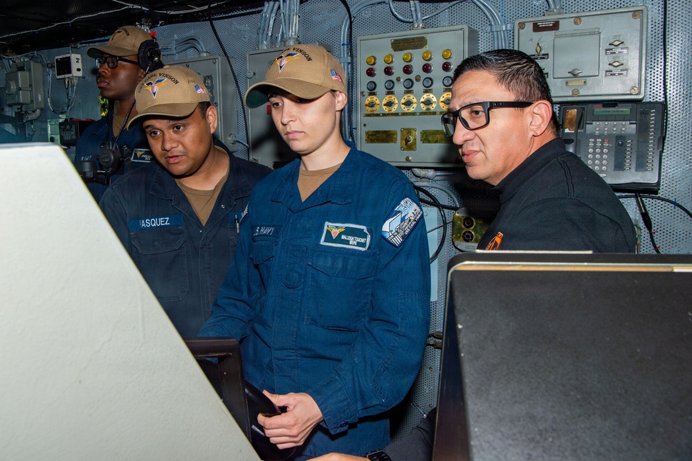 The image shows U.S. Navy personnel aboard what appears to be a naval vessel, likely USS Carl Vinson based on the visible cap insignia. Three to four individuals are visible in a ship's control or operations room, with one civilian or off-duty personnel wearing black clothing and glasses. The personnel are gathered around control panels and monitors in what appears to be a briefing or operational review.