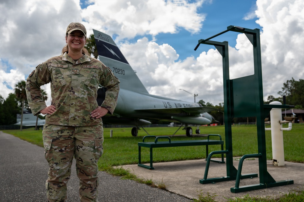 A U.S. Air Force service member wearing OCP (Operational Camouflage Pattern) uniform stands confidently with hands on hips in front of a static display aircraft. The aircraft visible in the background is a U.S. Air Force jet with tail number 70230 and 'Florida' markings, consistent with a gate guard or museum display. The setting appears to be an outdoor fitness or recreational area on a military installation.