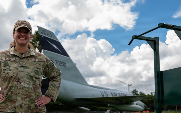 A U.S. Air Force service member wearing OCP (Operational Camouflage Pattern) uniform stands confidently with hands on hips in front of a static display aircraft. The aircraft visible in the background is a U.S. Air Force jet with tail number 70230 and 'Florida' markings, consistent with a gate guard or museum display. The setting appears to be an outdoor fitness or recreational area on a military installation.