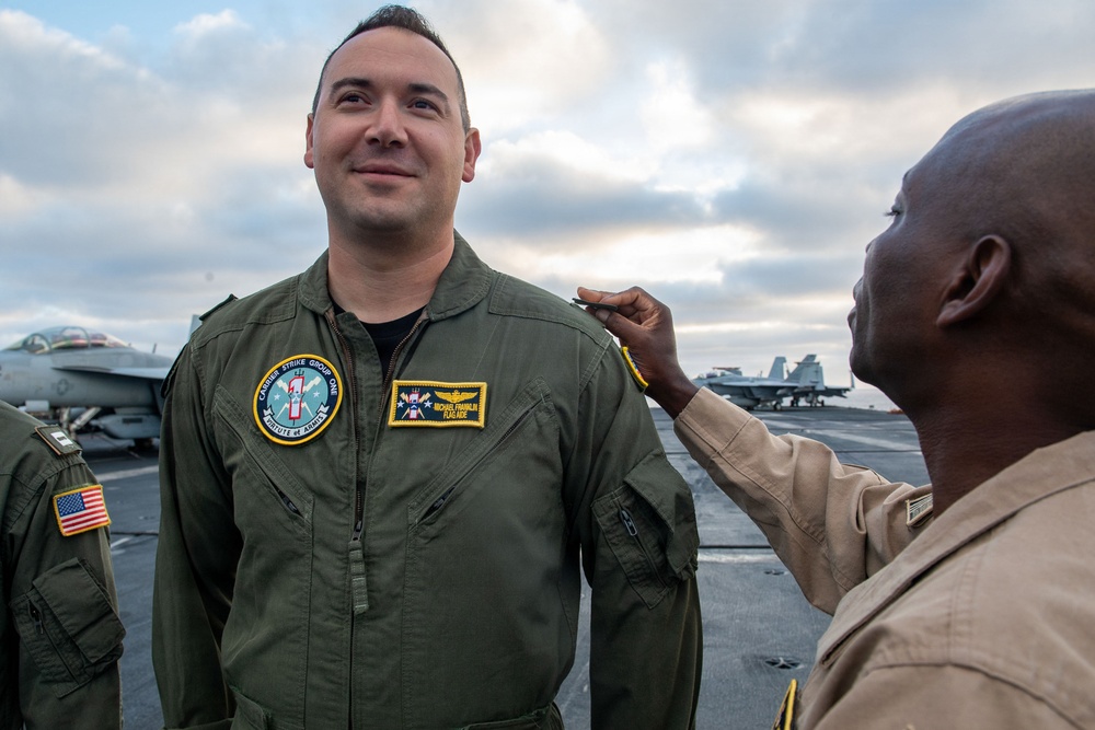 A military ceremony appears to be taking place on the flight deck of a U.S. Navy aircraft carrier, where one service member is pinning or adjusting insignia on another's uniform. The central figure wears a green flight suit with a 'Carrier Strike Group One' patch and a name tag reading 'Michael Franklin' with the title 'Flag Aide.' Military aircraft are visible in the background on the flight deck.