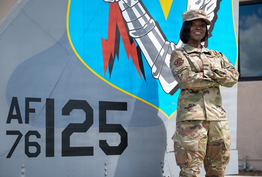 A U.S. Air Force service member in Operational Camouflage Pattern (OCP) uniform stands with arms crossed in front of a military aircraft tail section. The aircraft displays the tail code 'AF 76-125' and a unit emblem featuring a fist holding a lightning bolt on a blue shield background. The subject is smiling and posed in a confident stance for what appears to be an official or promotional photograph.