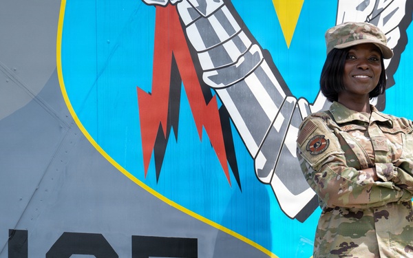 A U.S. Air Force service member in Operational Camouflage Pattern (OCP) uniform stands with arms crossed in front of a military aircraft tail section. The aircraft displays the tail code 'AF 76-125' and a unit emblem featuring a fist holding a lightning bolt on a blue shield background. The subject is smiling and posed in a confident stance for what appears to be an official or promotional photograph.