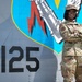 A U.S. Air Force service member in Operational Camouflage Pattern (OCP) uniform stands with arms crossed in front of a military aircraft tail section. The aircraft displays the tail code 'AF 76-125' and a unit emblem featuring a fist holding a lightning bolt on a blue shield background. The subject is smiling and posed in a confident stance for what appears to be an official or promotional photograph.