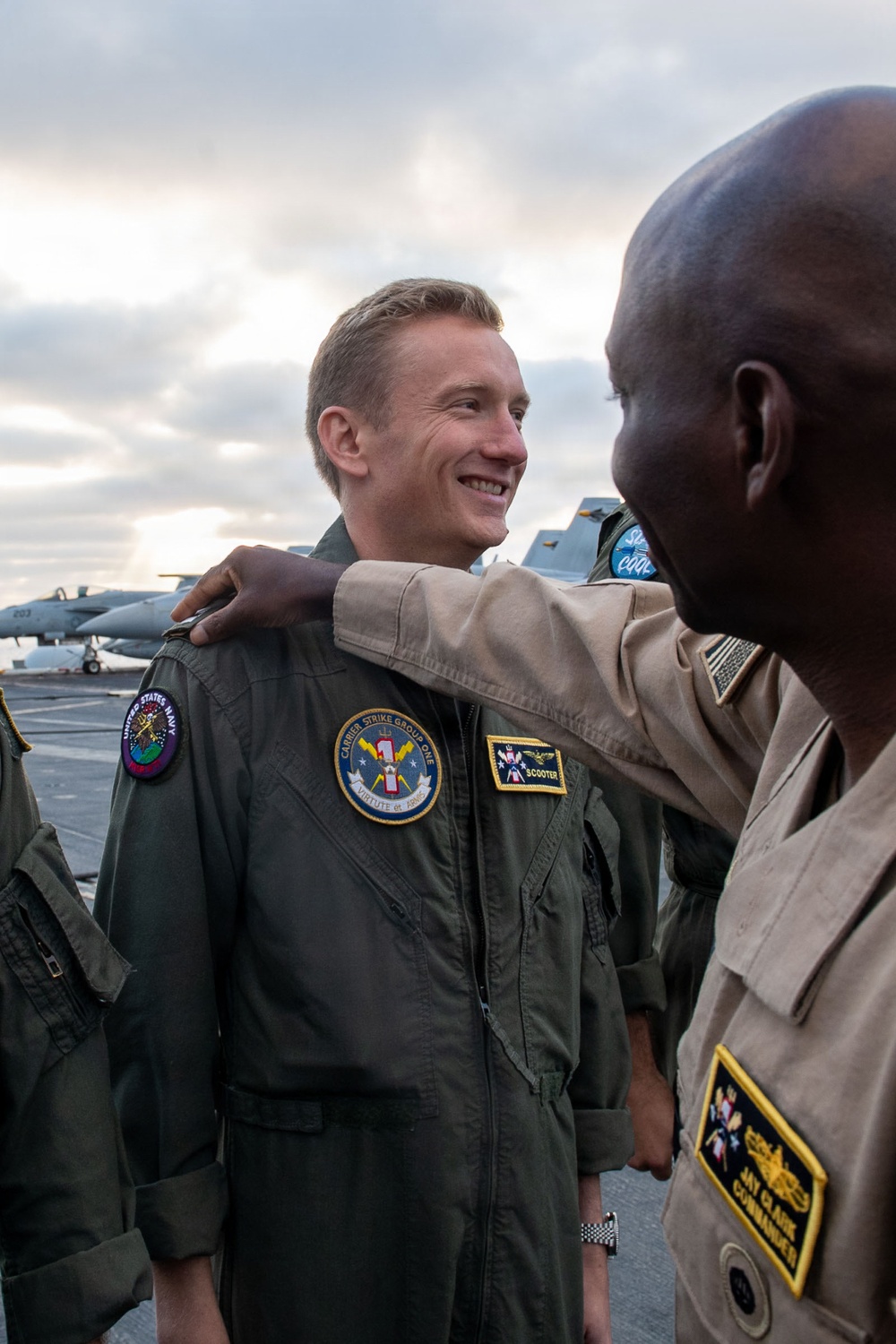 Two U.S. Navy personnel are interacting on what appears to be an aircraft carrier flight deck. The central figure, wearing a green flight suit with callsign 'SCOOTER' and a 'Carrier Strike Group One' patch, is smiling while a second individual identified by nameplate as 'JAY CLARK, Commander' places a hand on his shoulder. Military aircraft are visible in the background.