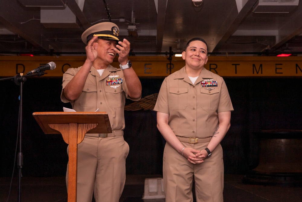 Two U.S. Navy personnel in khaki service uniforms stand together during what appears to be a formal military ceremony, likely a promotion or award event. A senior officer stands at a wooden podium with a microphone, gesturing and smiling, while a second service member stands at attention beside him with hands clasped. The setting appears to be aboard a naval vessel, indicated by the industrial overhead structure and a yellow banner reading 'DEPARTMENT' in the background.