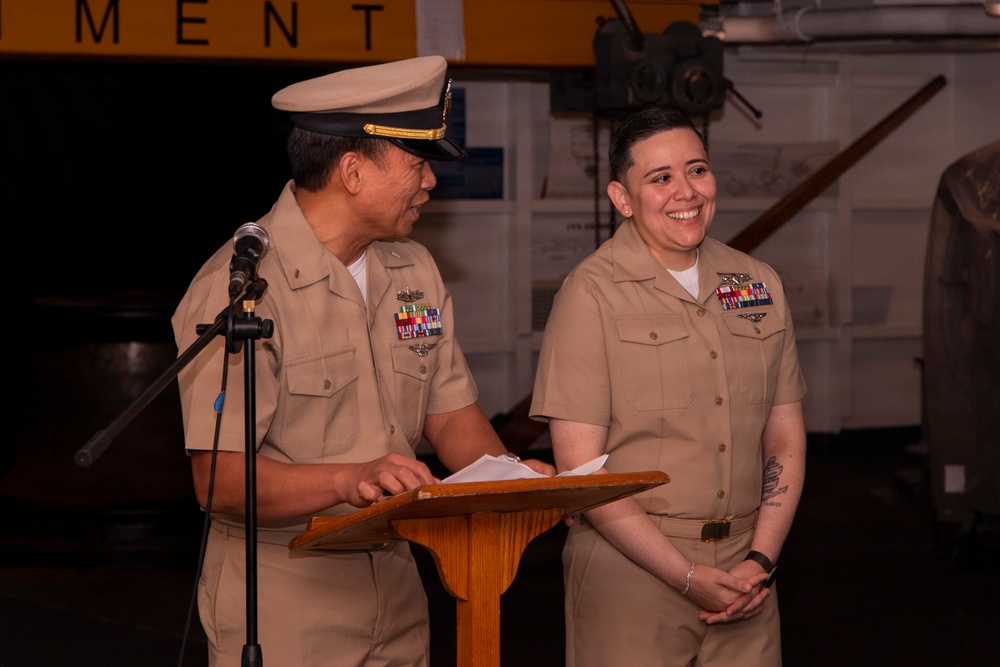 Two U.S. Navy personnel in tan service uniforms are standing at a wooden podium during what appears to be a formal military ceremony. The senior officer on the left is speaking at a microphone while the other service member stands beside him smiling. The setting appears to be inside a military facility or museum, with yellow signage visible in the background.