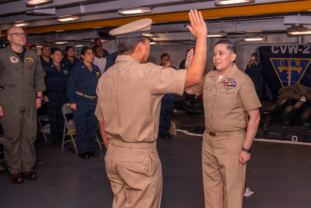 A military ceremony is taking place aboard what appears to be a U.S. Navy aircraft carrier, identifiable by the 'CVW-2' insignia visible in the background. Two naval personnel in tan Service Khaki uniforms are raising their right hands, consistent with an oath of office or re-enlistment ceremony. A group of uniformed military personnel stand as witnesses in the background.
