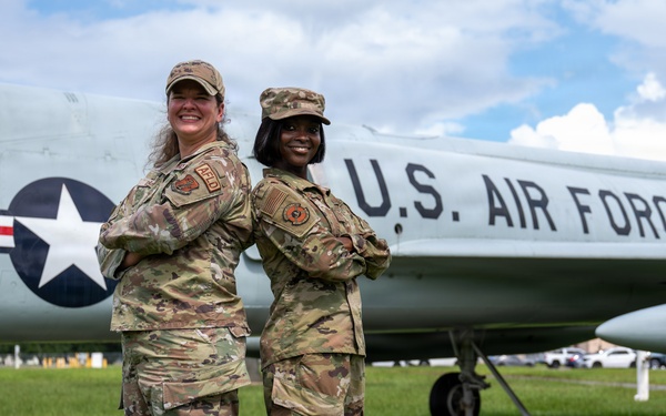 Two female U.S. Air Force personnel in OCP (Operational Camouflage Pattern) uniforms pose back-to-back with arms crossed in front of a static display aircraft marked 'U.S. AIR FORCE'. The aircraft appears to be a vintage jet, likely a B-57 or similar era aircraft, displayed on a grass field. Both individuals are smiling and appear to be in a formal yet celebratory pose.