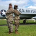 Two female U.S. Air Force personnel in OCP (Operational Camouflage Pattern) uniforms pose back-to-back with arms crossed in front of a static display aircraft marked 'U.S. AIR FORCE'. The aircraft appears to be a vintage jet, likely a B-57 or similar era aircraft, displayed on a grass field. Both individuals are smiling and appear to be in a formal yet celebratory pose.